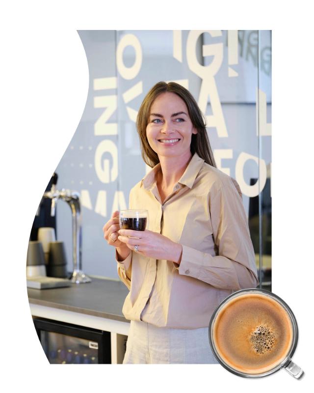 Smilende dame står op ad et bord med en kop kaffe i hænderne og kigger til venstre