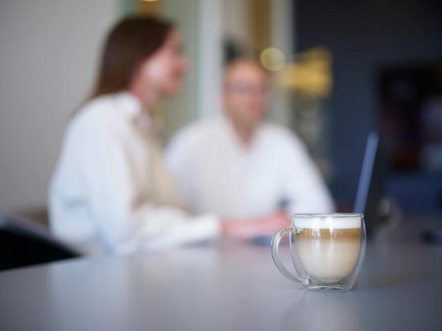 Billede af et glas med kaffe i på et bord og to mennesker sløret i baggrunden ved en computer